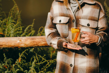 Woman in checked coat holding glass cup of delicious white mulled wine with a cinnamon stick in her hands. Warming drink in autumn.