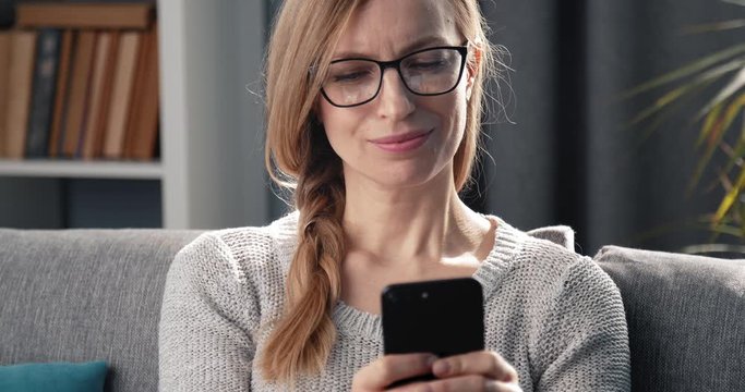 Portrait of happy woman in home clothing and eyeglasses sitting on couch and texting on smartphone. Mature lady with blond hair sending messages while relaxing at home.