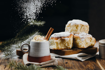 A hot cocoa with whipped cream, cinnamon stick and fresh Christmas buns with powder and over festive table, sugar powder is sprinkled on the background lke a snow. Winter holidays.