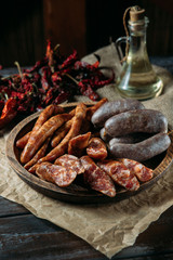 Traditional three kinds of meat sausages in rustic wood bowl, dried chili peppers and oil in a glass jug on natural black wood table.