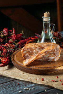 Large piece of traditional dish head cheese and its slice on wooden cutting board on craft paper, dried peppers on the backgrounds.
