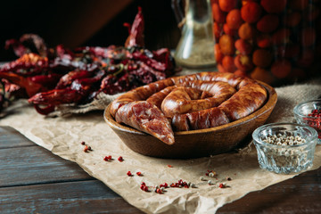 Horizontal photo of homemade raw chopped sausages in a wooden bowl, dried peppers and pickled tomatoes on the wooden table.