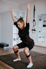 EMS training. Girl doing squats with her arms raised in suit with cables.