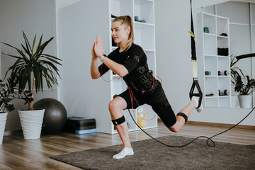 Woman doing lunges in EMS electro stimulation suit with cables, using TRX.