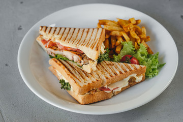 Plate with two chicken and bacon club sandwiches with fries and vegetables on a white plate on a concrete background.