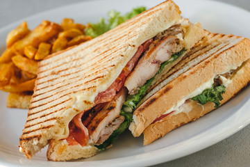 Plate with two chicken and bacon club sandwiches with fries and vegetables on a white plate on a concrete background.