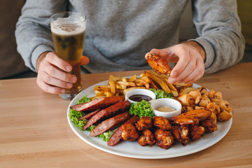 Man having dinner. Man's hands holding chicken leg and beer, large hot beer plate with sausages, fries, chicken and sauses on the table.