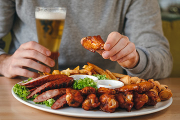 Man having dinner. Man's hands holding chicken leg and beer, large hot beer plate with sausages, fries, chicken and sauses on the table.