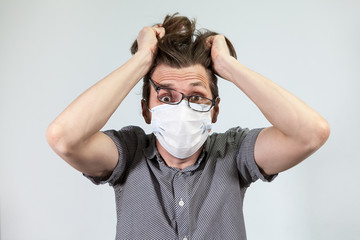 Scared Caucasian man in disposable mask and glasses tears his hair out, male in panic, grey background