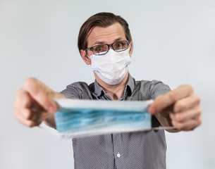 European man in glasses and one-use mask holds out a disposable medical mask, grey background