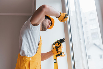 Handyman in yellow uniform installs new window by using automatic screwdriver. House renovation...