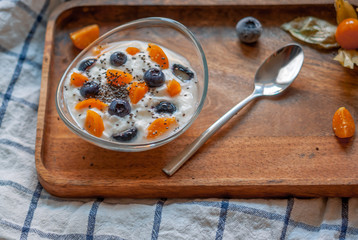 Breakfast yogurt in a glass bowl with blueberries and physalis on a wooden brown tray on a concrete gray-blue background with a towel in a thin blue check.