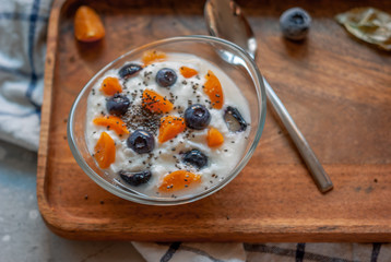 Breakfast yogurt in a glass bowl with blueberries and physalis on a wooden brown tray on a concrete gray-blue background with a towel in a thin blue check.