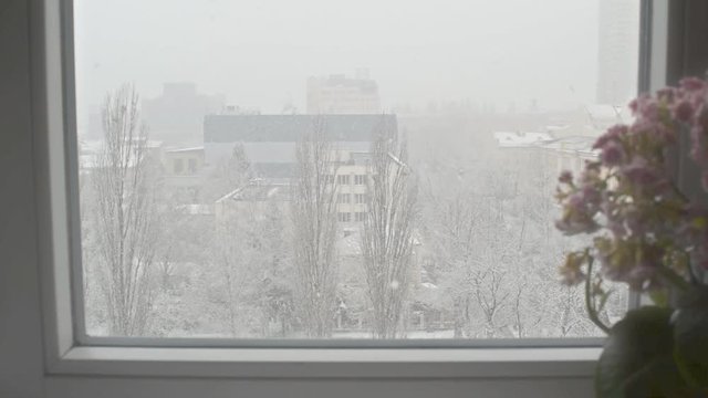 Unexpected snowfall outside the window of a city apartment. White fluffy snow covers the square, streets and houses of the cityscape. The arrival or return of winter. View from above.