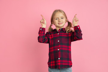 Pointing up, smiling. Caucasian little girl's portrait on pink studio background. Beautiful female model with blonde hair. Concept of human emotions, facial expression, sales, ad, youth, childhood.