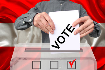 male voter drops a ballot in a transparent ballot box against the background of the national flag...
