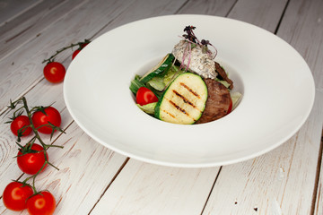 Grilled zucchini vegetable salad with a slice of marbled beef steak. A wooden table is decorated with a bunch of cherry tomatoes.