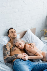 Attractive girl in underwear touching hand of muscular tattooed boyfriend on bed