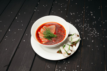 Tender borscht with meat served on a deep white plate with croutons and lard on a black wooden table sprinkled with sea salt