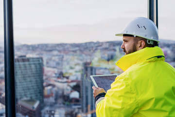Man engineer standing on construction site, holding tablet with blueprints.