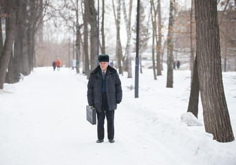 Fototapeta na wymiar An old man with suitcase standing outdoors in winter park while snowfall