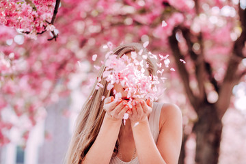 Closeup portrait of a lovely girl in a park with blooming japanese sakura trees. Romantic young blonde in a dress posing on a background of spring flowering trees. Flying cherry petals.