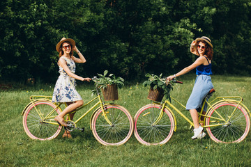 Two pretty girls go with bicycles in a spring park at sunset. Girlfriends have fun, laugh, smile and ride bicycles. Full-length portrait of two young women in dresses and straw hats. active holiday