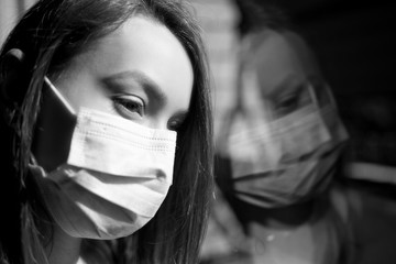 Girl in a mask looks out the window, in quarantine with reflection. Gloomy frame, quarantine. Powerful outbreak of coronavirus.