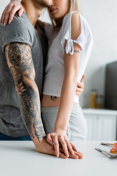 Cropped view of woman embracing tattooed boyfriend near kitchen table