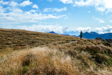 A woman in hiking outfit wandering in Italian Alps. She walks between golden grass. Hard to reach mountain peaks. There are many mountain ranges in the back. Serenity and calmness. Freshness