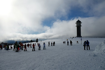 Zahlreiche Ski - und Snowboardfahrer am Feldberg im Schwarzwald. Oberer Bereich des schneebedeckten Skigebietes. Feldbergturm im Hintergrund zu sehen