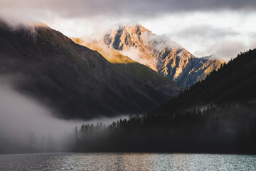 High gold mountain peak and many low clouds above mountain lake at sunrise. Dense fog above water and forest in golden hour. Atmospheric highland landscape at early morning. Alpine relaxing scenery.