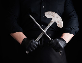 male cook in black uniform and black latex gloves holds a large sharp vintage knife for meat and vegetables and a metal sharpener for knives