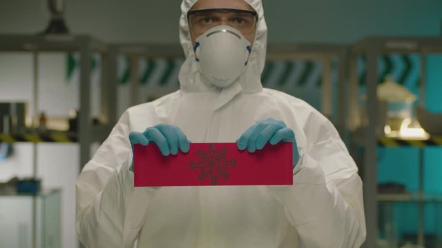 VICTORY OVER THE VIRUS. Portrait of masked laborant . Concept of virus quarantine. Doctor with a overall safety uniform Tear the red paper with the virus cell. Shot on ARRI ALEXA camera in slow motion