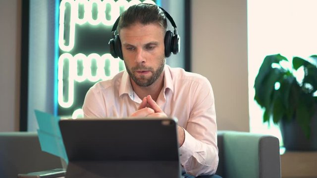 Smart casual man in electronic headphones for noise cancellation having online seminar on modern touch pad for web teaching, millennial male in bluetooth equipment talking during video call