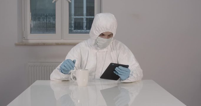 Portrait of young male person stitting behind white dining desk in kitchen in protective suit with a cup of tea and reading news on his tablet during pandemic coronavirus quarantine time 4K