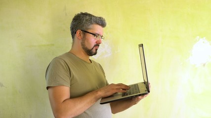 A man with glasses and a beard on his face. Holds a laptop in his hand. Types on the keyboard. Against the background of a green wall. Half-length portrait. Builder at the facility. Freelancer at home