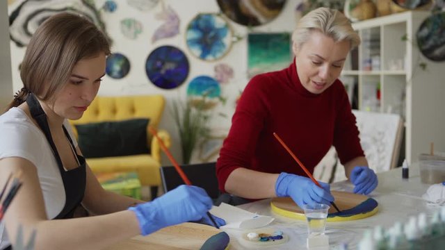 Tracking medium shot of two middle aged female artists painting with blue acrylic paints on wooden round canvases and talking sitting together at desk in art studio