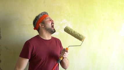 Bearded man against the wall. Sings in a construction roller. Performance of the artist. Musical number. On the head is an orange ribbon. Repair work in the room. Frick. Retro style. Green wall. Rock-