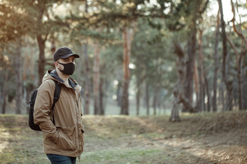 Young man in medical mask standing in park. Man with backpack in jacket and black cap 