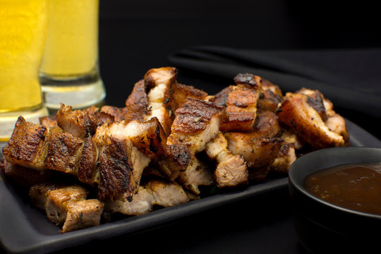 Grilled pork belly in a black platter with barbecue salce and beer in black background