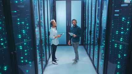 Couple of mixed-race technicians walking in database corridor of server room discussing work solving tech issues coworking in data center cyber security.
