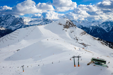 Fototapeta na wymiar Landscape at a ski resort Campitello di Fassa Italy. Winter Dolomites and blue sky with clouds. Aerial view on ski slopes and ski lifts