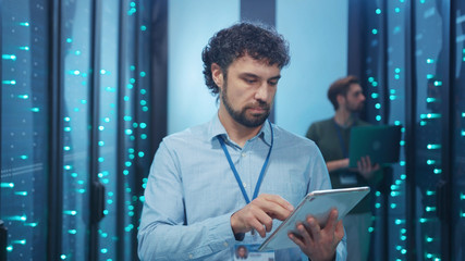 Data center specialist working on digital tablet in high tech server room. Handsome cheerful technician smiling at camera coworking in computer security.