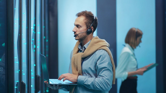 IT professional in server room visually inspecting secure data center cages running diagnostics on computer laptop coworking with female colleague.
