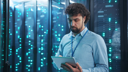 Bearded head administrator running backups on digital tablet in rack server room. His young...