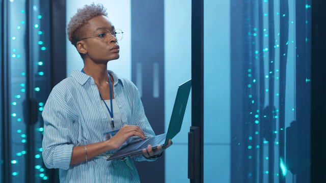 Professional african woman administrator running data center backups operation on open server rack cabinet working in digital high tech internet room.
