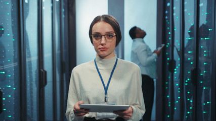 Beautiful woman IT administrator holding tablet looking at camera cooperating with african colleague in server room of data center. Cyber security.