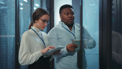 Data center black chief administrator teaching young female intern inspecting working rack servers...
