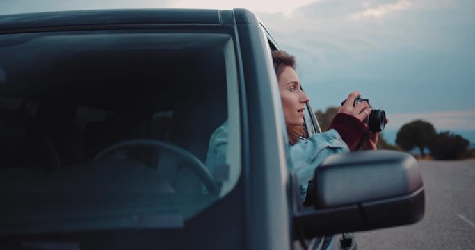 Young hipster girl traveling in car exploring Europe, attractive female traveller taking photos of sunset on camera while sitting in an off-road car, slow motion